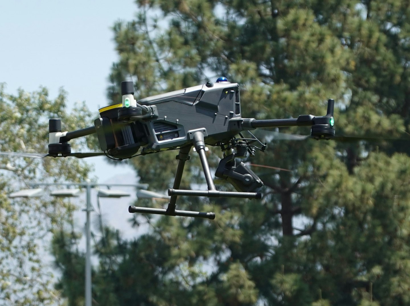 A grey and black drone hovering against a blue sky.