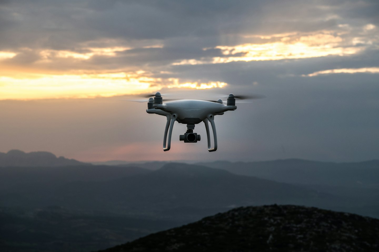 A white drone flies over mountains at sunset.